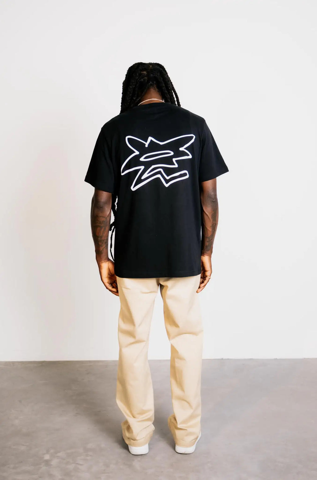 Full-body back view of a model in studio wearing the grazzy Symbol graphic t-shirt in the Black colorway, featuring the big white flower-like grazzy logo mark on the back and showcasing the t-shirt's relaxed fit and silhouette.