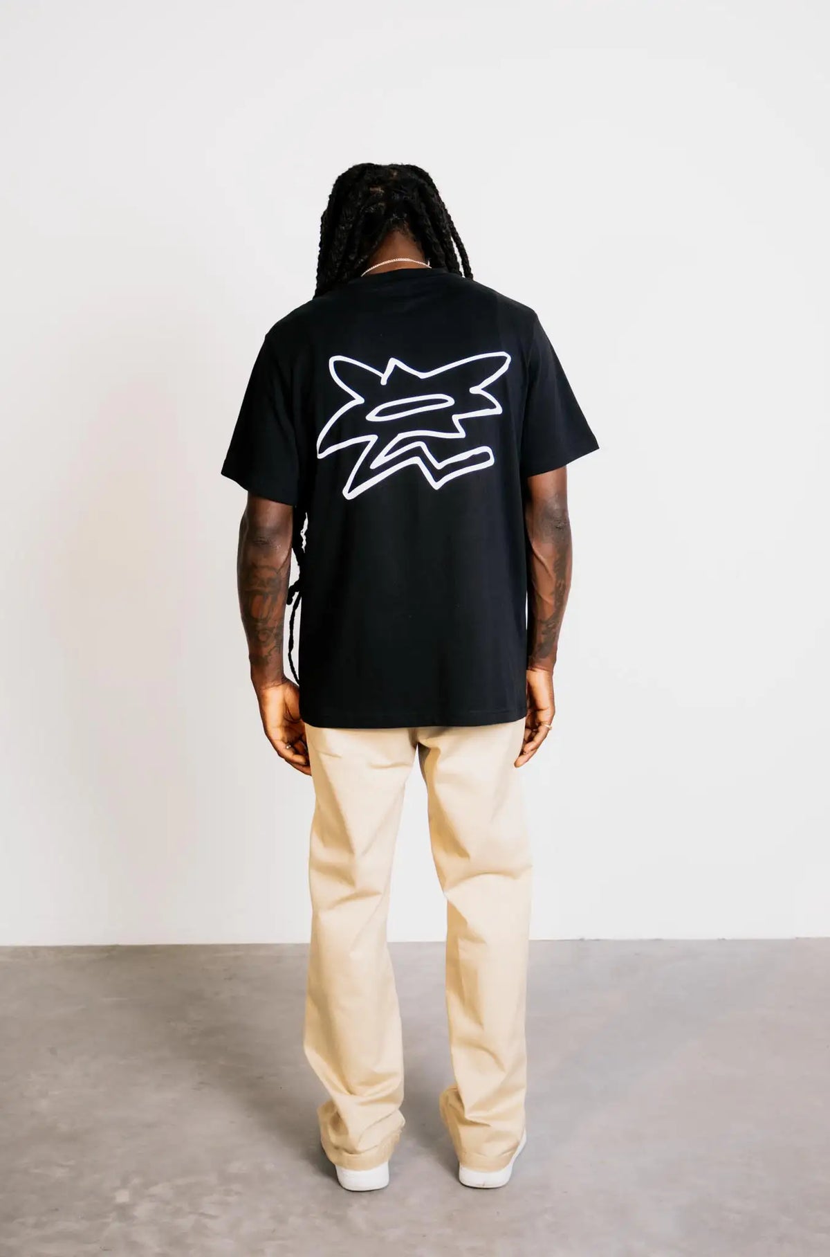 Full-body back view of a model in studio wearing the grazzy Symbol graphic t-shirt in the Black colorway, featuring the big white flower-like grazzy logo mark on the back and showcasing the t-shirt's relaxed fit and silhouette.