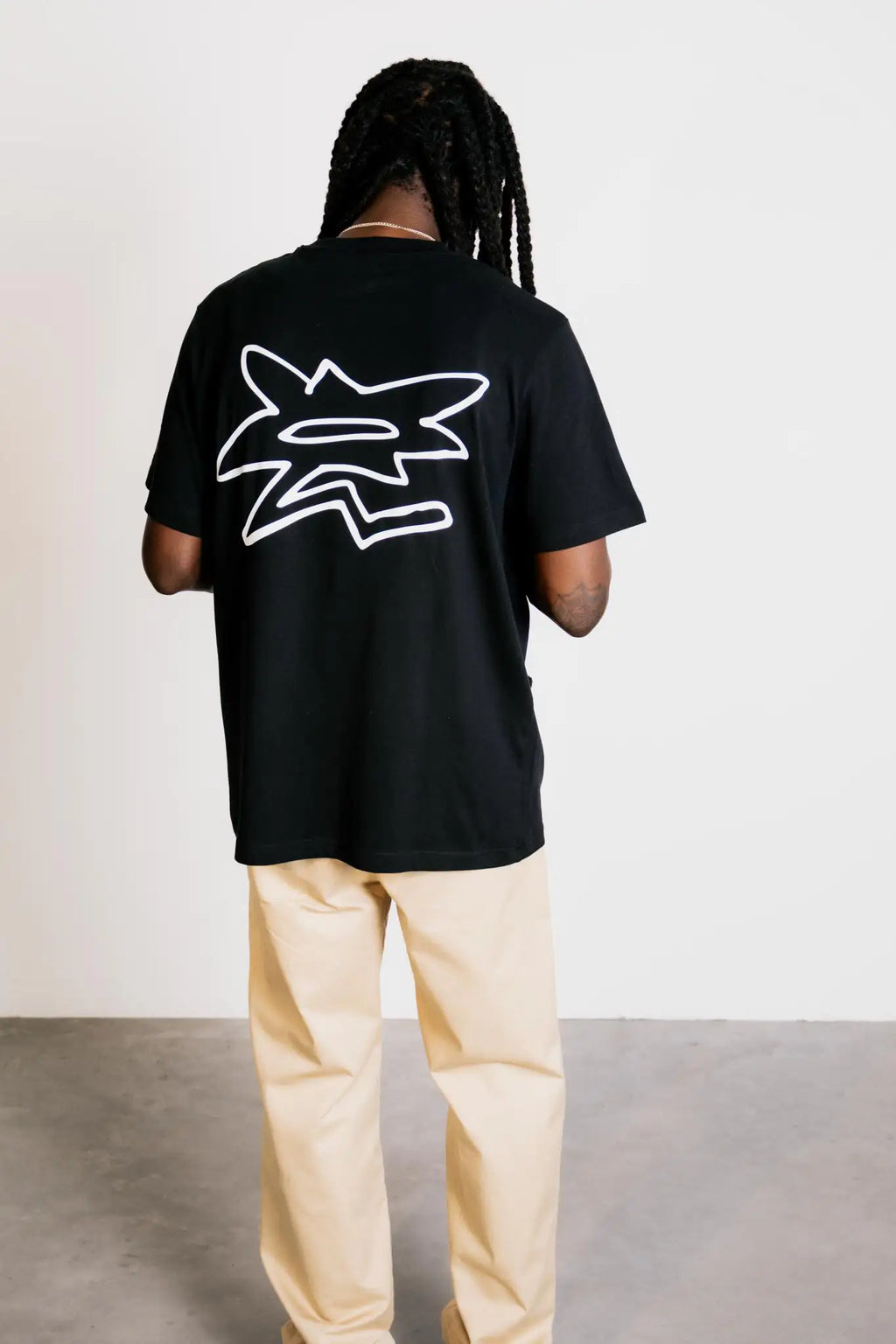 Full-body back view of a model in studio wearing the grazzy Symbol graphic t-shirt in the Black colorway, featuring the big white flower-like grazzy logo mark on the back and showcasing the t-shirt's relaxed fit and silhouette.