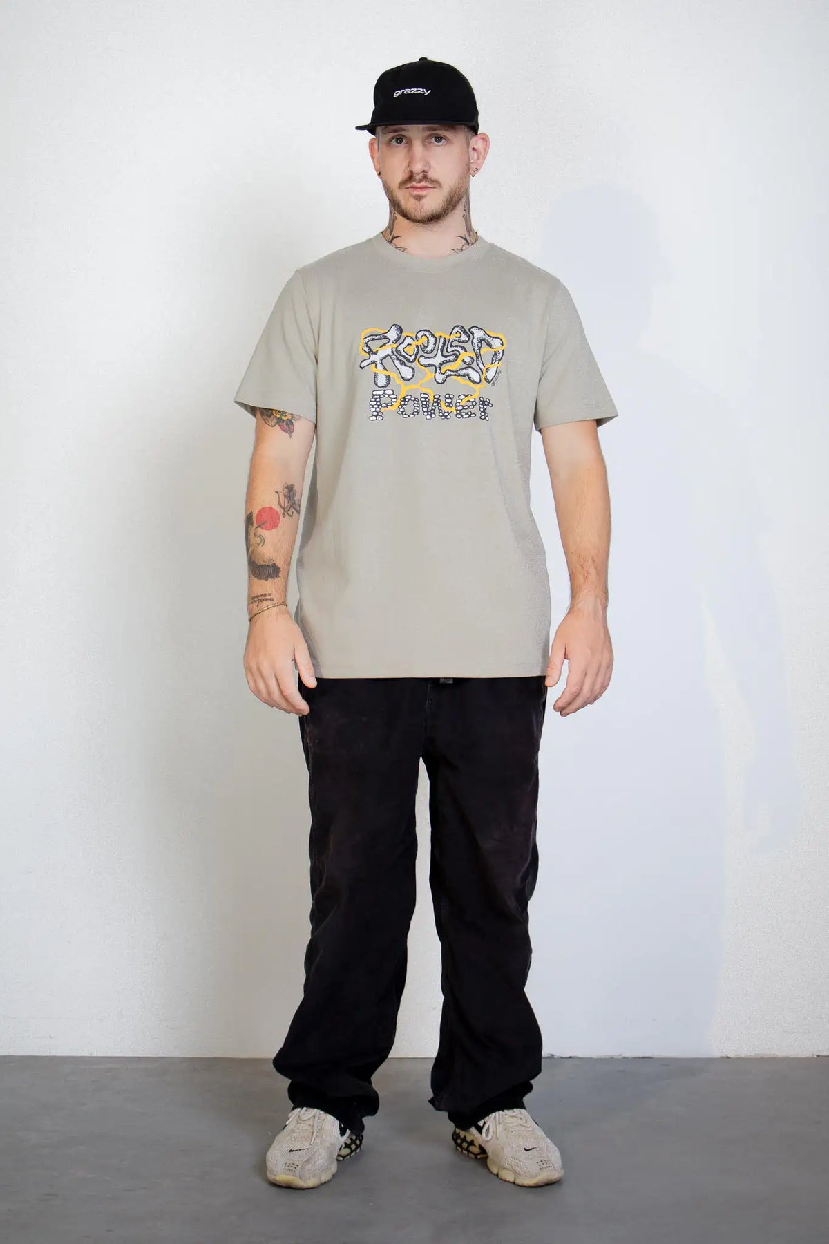 Full-body shotof a model in studio wearing the grazzy Rooted Power t-shirt in the Stone colorway and the grazzy Classic Logo black cap.